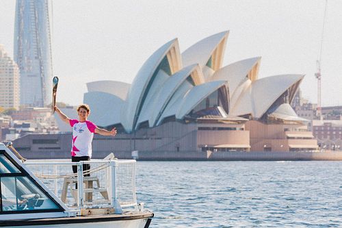 Australian diver Sam Fricker stands with the Queen's Baton at the Sydney Harbour, with the Sydney Opera House directly behind him.