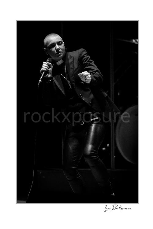 Vertical black and white image of Sinéad O’Connor singing into a microphone in a dramatic low-light stage portrait