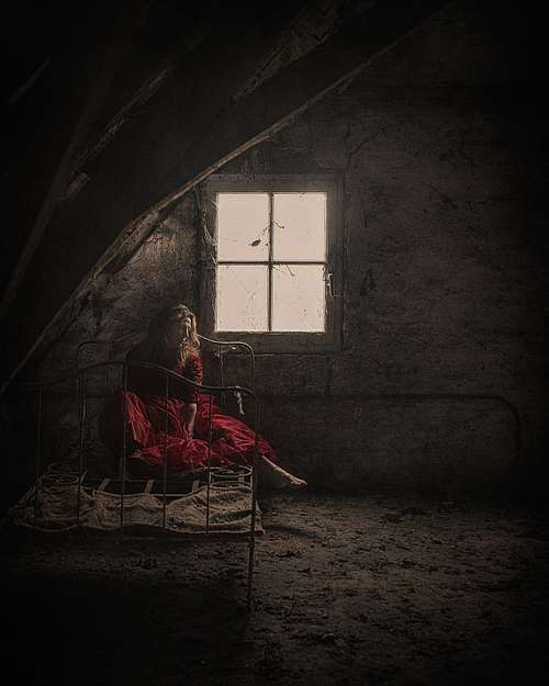 Portrait artistique d'une femme en robe rouge assise sur un lit ancien dans un grenier sombre, &eacute;voquant l&rsquo;abandon, la solitude et la reconstruction int&eacute;rieure &mdash; photographie fine art par Pascal Yennis, Namur, Belgique.