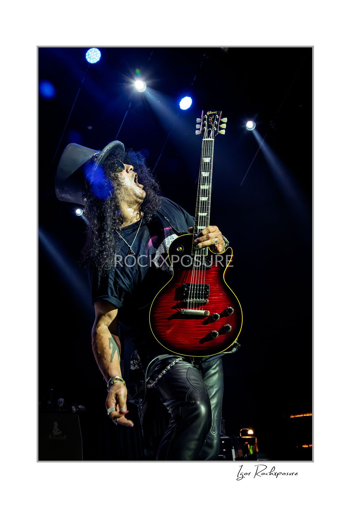 Vertical concert photography of Slash holding his electric guitar raised beside him wearing his signature top hat under blue stage lighting