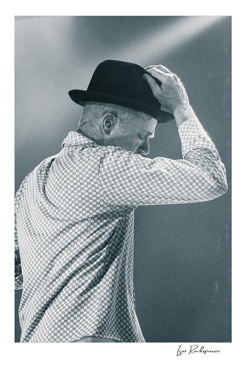 Vertical black and white image of Gord Downie of The Tragically Hip in a quiet live performance moment, touching the brim of his hat under soft stage light