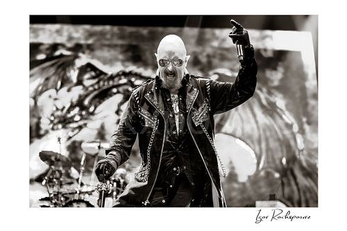 Horizontal black and white image of Rob Halford of Judas Priest performing live on stage, holding a microphone and cane in front of a dramatic LED screen backdrop