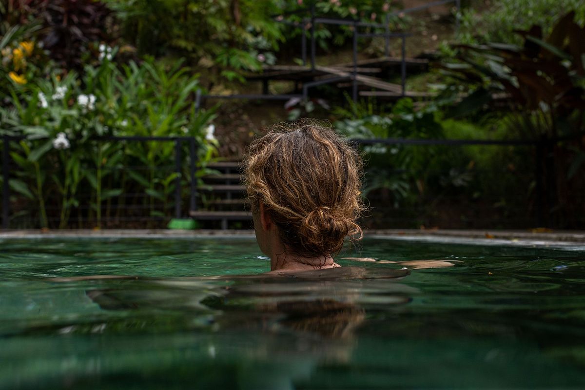 Redaktionellt milj&ouml;portr&auml;tt bakifr&aring;n: kvinna i pool i lugn tropisk milj&ouml; med gr&ouml;nska i bakgrunden.