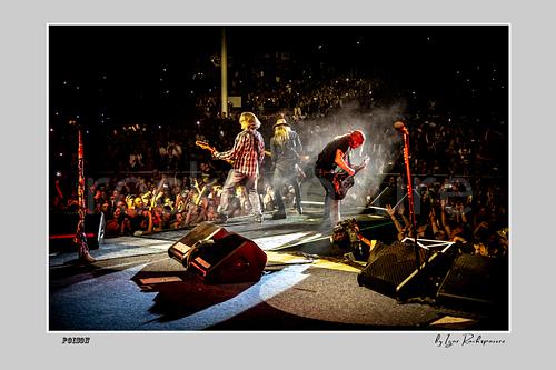 Horizontal color image of Bobby Dall, C.C. DeVille and Bret Michaels performing on a Poison stage, photographed with a remote camera mounted on the drum kit facing forward, with crowd lights and stage smoke