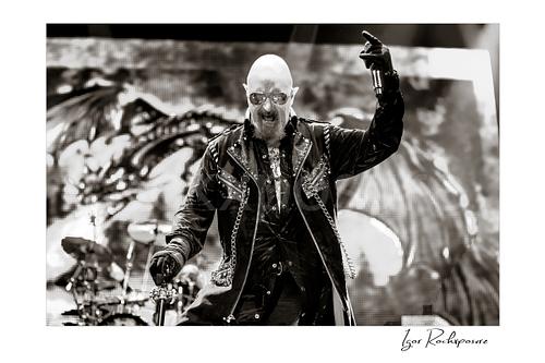 Horizontal black and white image of Rob Halford of Judas Priest performing live on stage, holding a microphone and cane in front of a dramatic LED screen backdrop