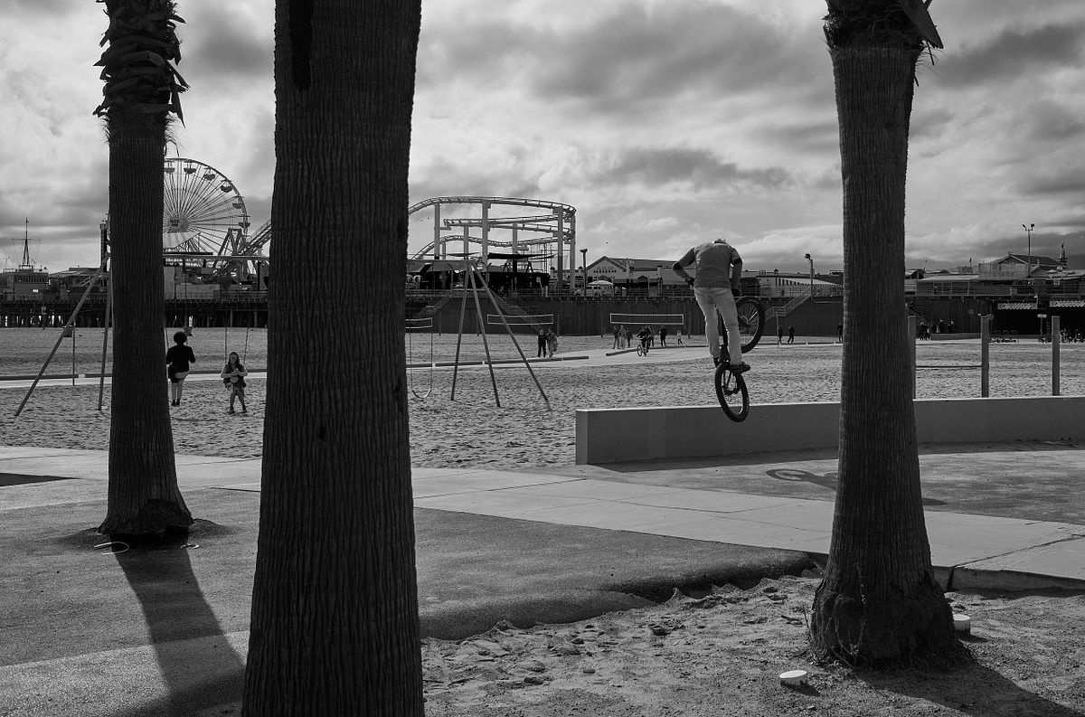 A BMX rider performing a stunt at a beach with palm trees and a ferris wheel in the background, captured by photographer Sandeep Gajula