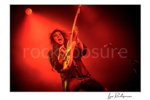 Horizontal color image of Yngwie Malmsteen performing live with a white guitar in a dramatic red-lit stage scene, hair flying and mouth open under dense concert haze