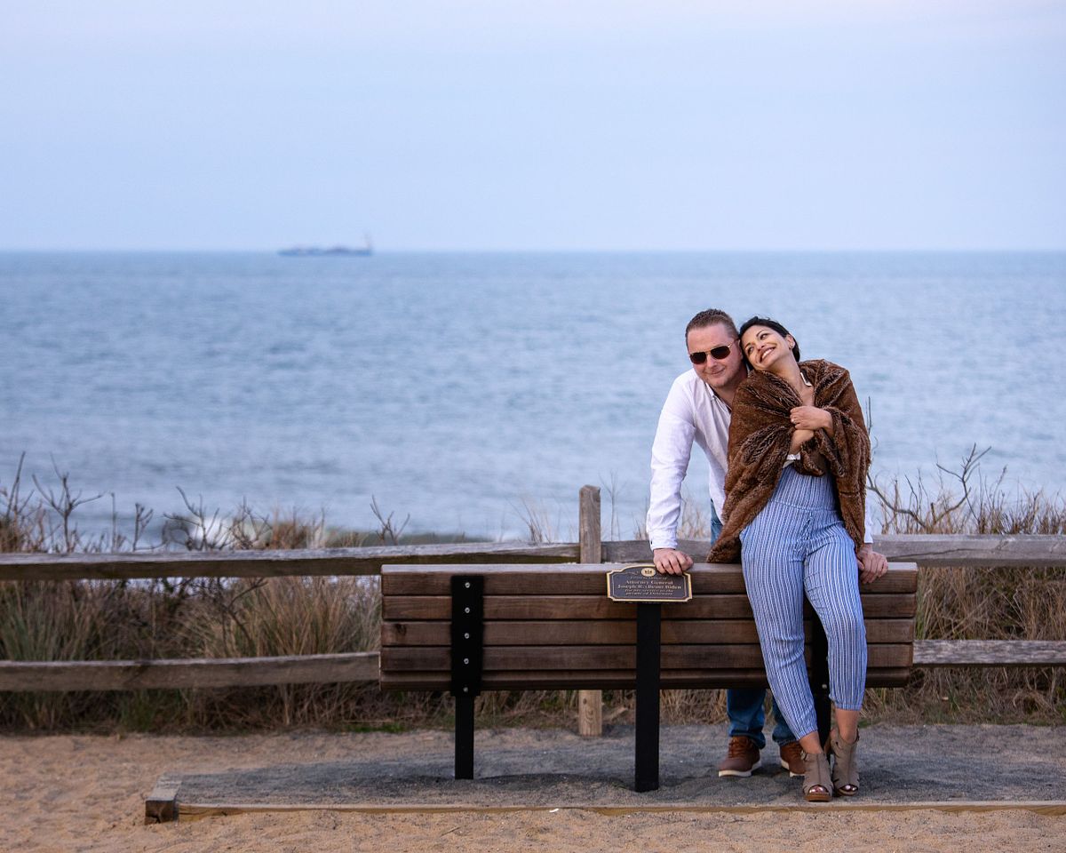 couple on cape henlopen on the biden memorial bench, she is in the front he is in the back. she is wearing a stole, the blue ocean is in the background