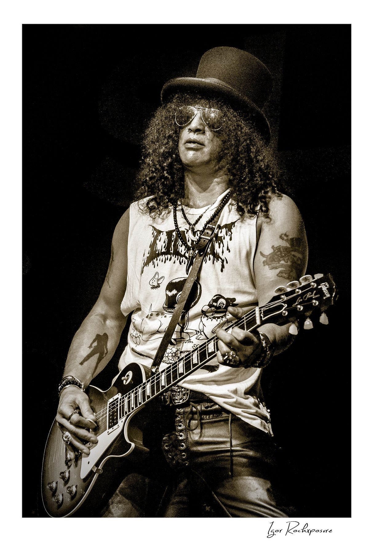 Vertical concert photography of Slash playing electric guitar wearing his signature top hat in black and white photography under stage lighting