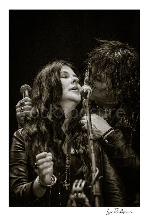 Vertical black and white image of Tom Keifer and Savannah sharing the microphone in a close live performance moment, framed tightly together at center stage