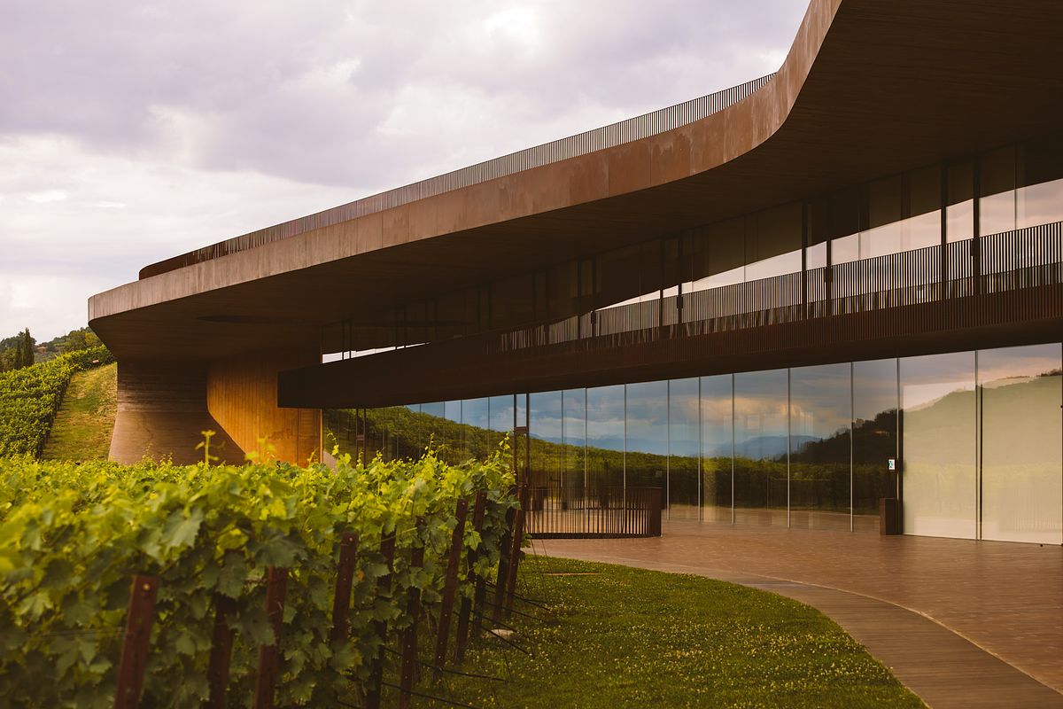 A wide exterior view of the Antinori winery in Tuscany, showcasing the sleek, horizontal Cor-Ten steel structure integrated into a lush green hillside planted with grapevines.