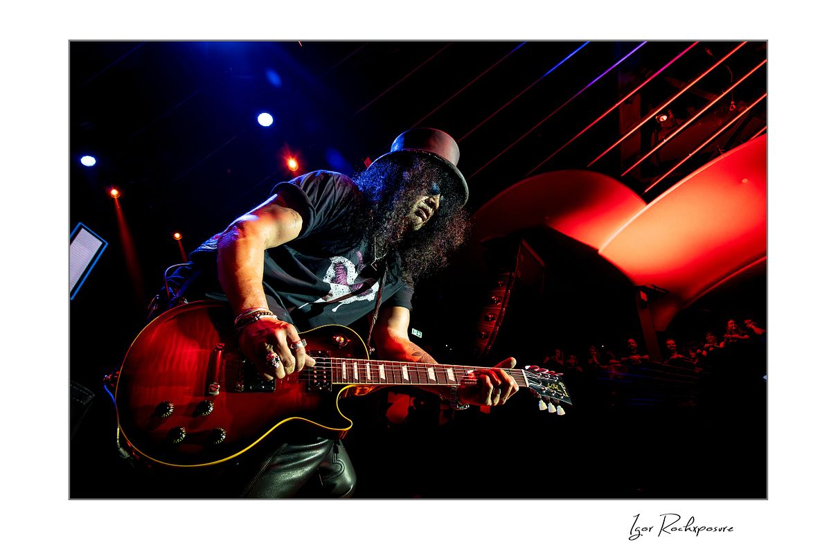 Horizontal concert photography of Slash playing electric guitar from a low angle under dramatic red and blue stage lighting