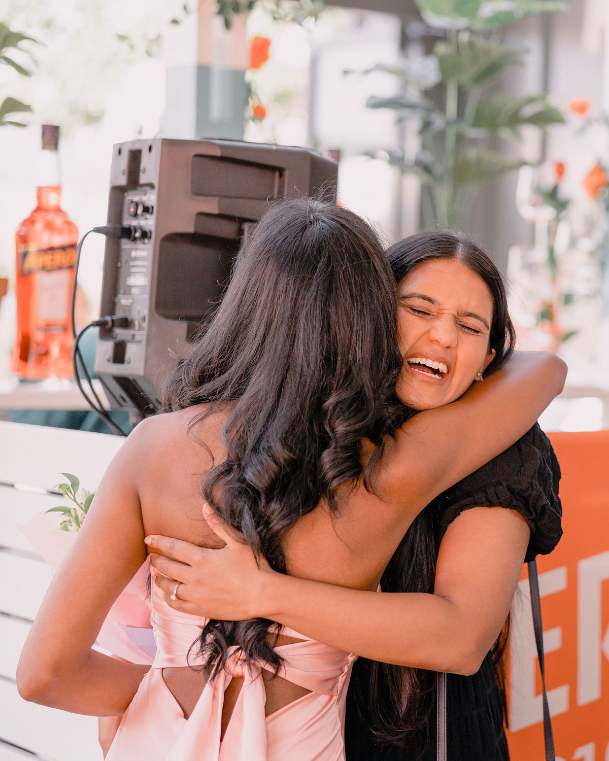 Photographer in Basel capturing two women at an engagement party are embracing joyfully; one in a pink dress holds flowers, the other in black is smiling widely. Background includes plants and a speaker.
