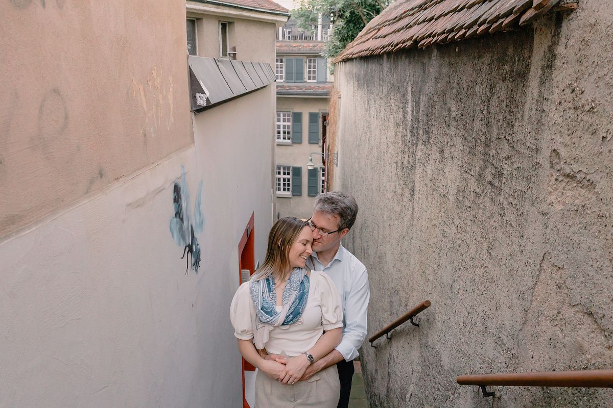 A Basel couple embraces lovingly on narrow outdoor stairs between aged walls. A blue butterfly graffiti is on the wall, and the scene feels intimate and warm.