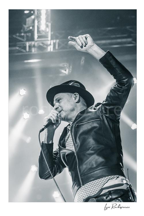 Vertical black and white image of Gord Downie of The Tragically Hip singing into a microphone with one arm raised, wearing a hat and leather jacket under bright stage lights