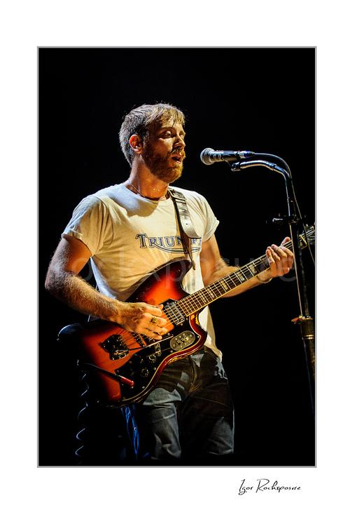 Vertical color image of Dan Auerbach of The Black Keys singing into a microphone while playing guitar under warm stage lighting