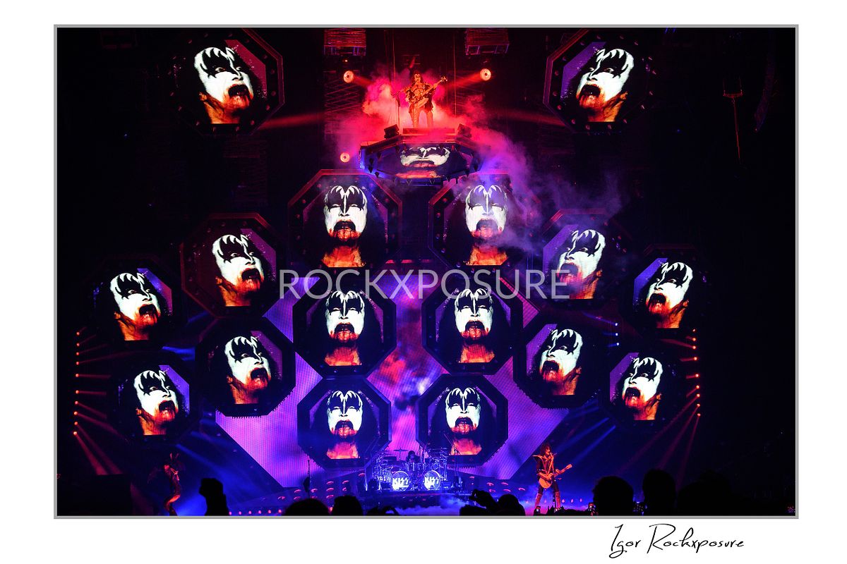 Gene Simmons of KISS standing on an elevated stage platform surrounded by large illuminated Gene Simmons demon face visuals across the stage screens under red and purple concert lighting