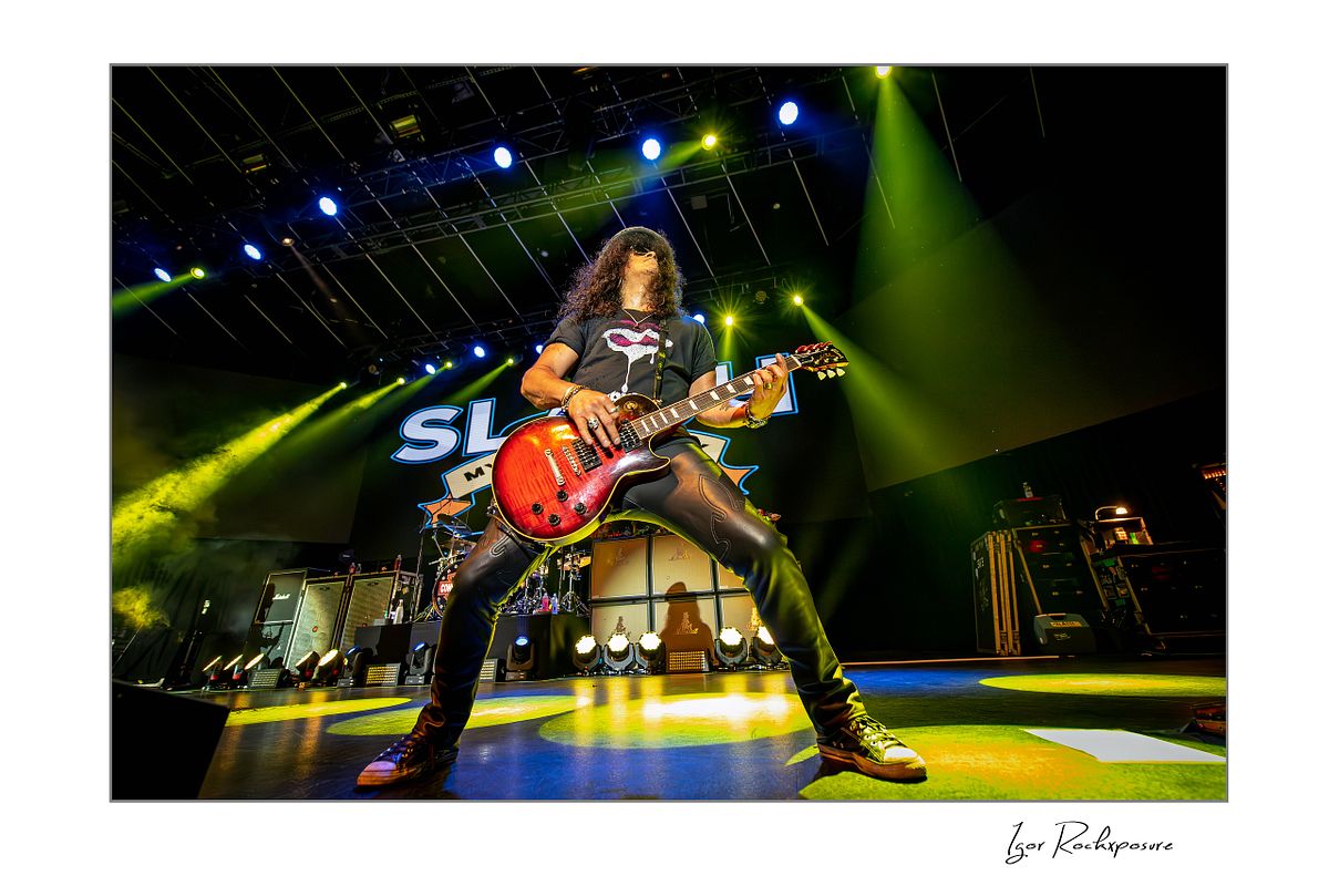 Horizontal concert photography of Slash playing electric guitar in a wide power stance under bright green and yellow stage lighting