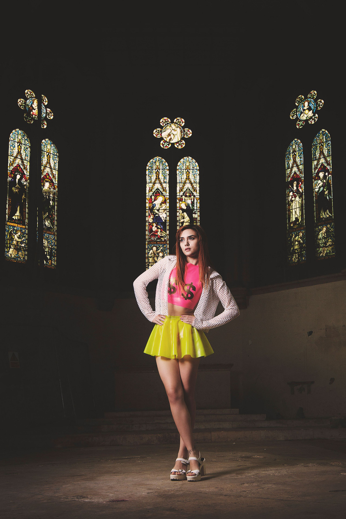 Full-length alternative fashion portrait of a model standing in a dark church interior with towering stained glass windows, captured by Carl Hewlett for Obsidian Noir. She is wearing a neon yellow latex skater skirt, a pink crop top with dollar signs by Shokushu Boutique, and a white mesh jacket.