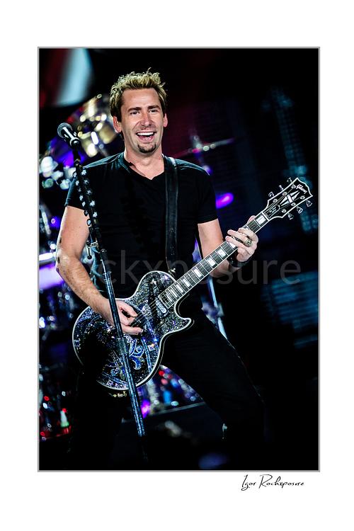 Vertical color image of Chad Kroeger of Nickelback playing guitar onstage beside a microphone stand with the drummer visible behind him