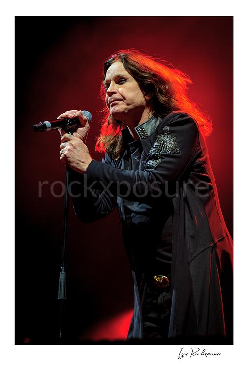 Vertical concert photography of Ozzy Osbourne of Black Sabbath singing into a microphone under dramatic red stage lighting
