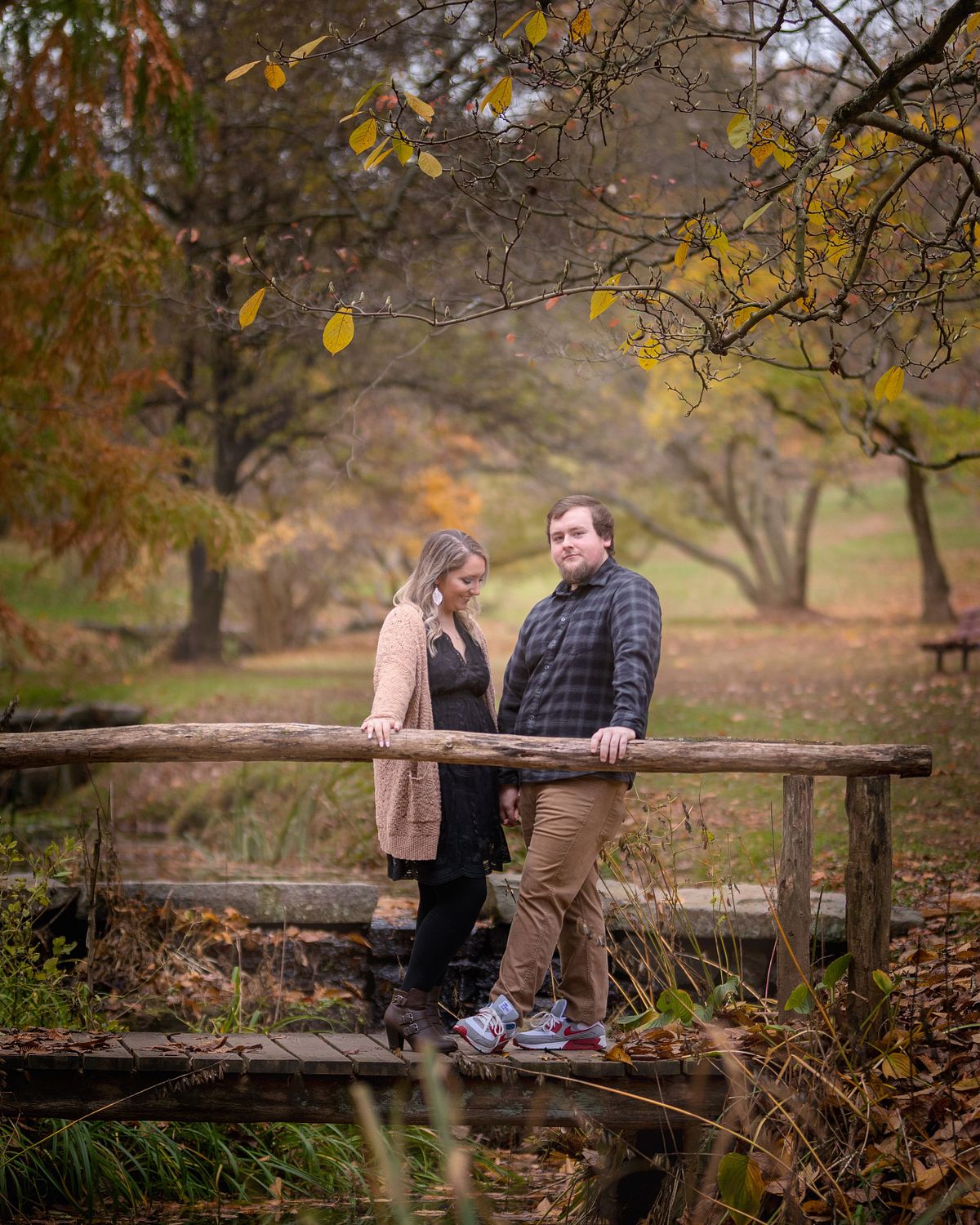 engagement photo during autumn with colourful leaves in the background. they are standing on a wooden bridge.