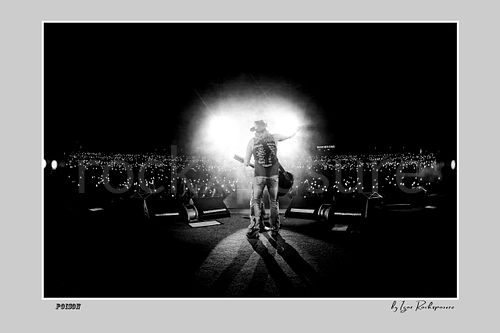 Horizontal black and white image of Bret Michaels on a Poison stage facing a sea of crowd lights with powerful backlight and long shadows