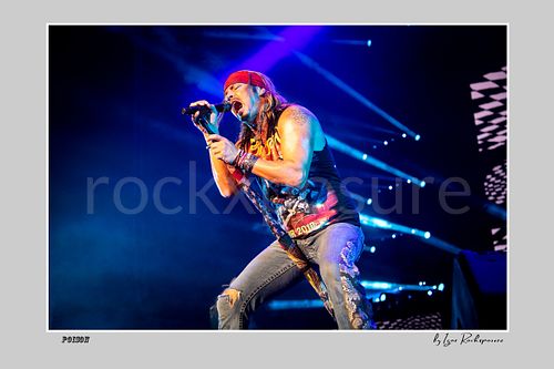Horizontal color image of Bret Michaels singing into a microphone during a Poison concert under vivid blue stage light