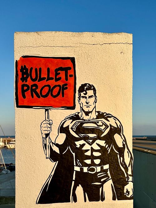 Street art by Street Cyber featuring Superman holding a red sign with "BULLET-PROOF" and a Bitcoin symbol. The artwork, in stencil style, symbolizes Bitcoin's resilience and strength against traditional financial vulnerabilities. This piece represents financial freedom, decentralization, and the power of digital assets. Located in Barcelona, it's a bold urban commentary. #Bitcoin #Bulletproof #Superman #StreetArt #FinancialFreedom #Decentralization #StreetCyberArt #ArtForChange #UrbanArt #Barcelona