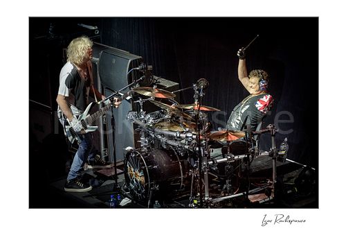 Horizontal color image of Rick Savage and Rick Allen of Def Leppard performing live on stage, with Savage at left playing bass guitar and Allen at right behind the drum kit holding a drumstick aloft